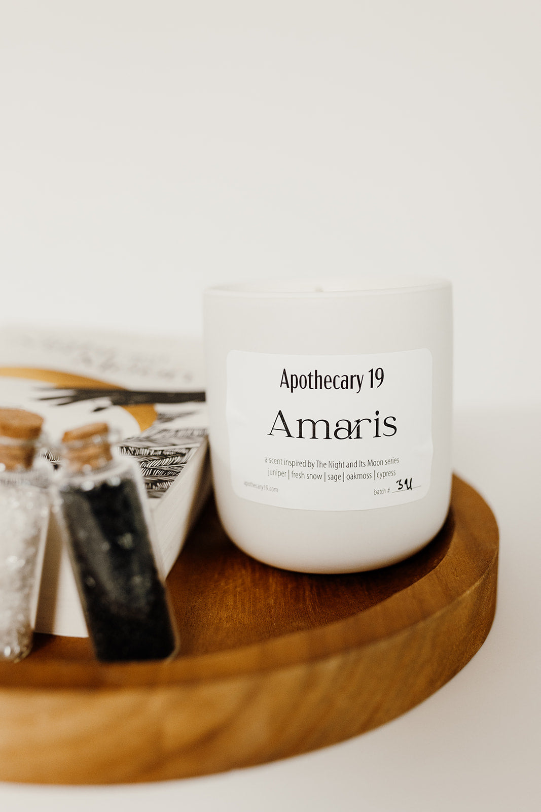 white ceramic jar candle sitting on a wooden plate. There are two small vials of white and black crystals and a book titled, The Night and Its Moon