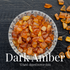 A glass bowl of amber-coloured tumbled stones on dark textured stone, with more amber pieces scattered around it, labeled Dark Amber in white